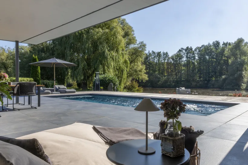 Blick von der überdachten Terrasse über das Becken auf Teich und Trauerweide