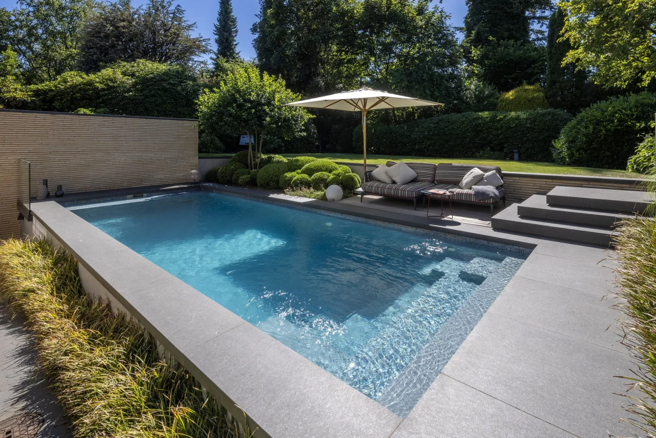 Becken mit Natursteinrand, Sonnenschirm und Lounge am Rasen