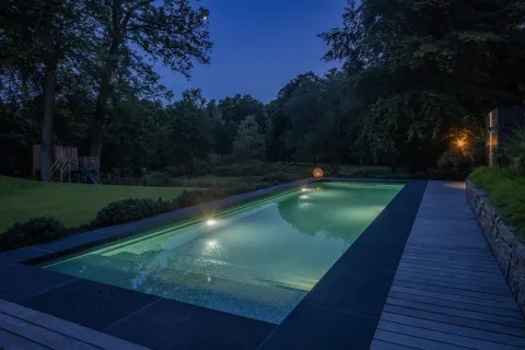Beleuchtetes Becken in der Dämmerung, dahinter Garten und Waldrand