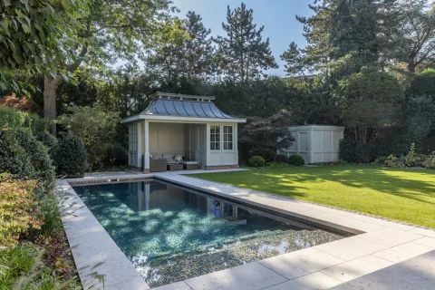 Becken mit schimmerndem Mosaik vor einem Poolhaus mit Metalldach, alter Baumbestand ringsum