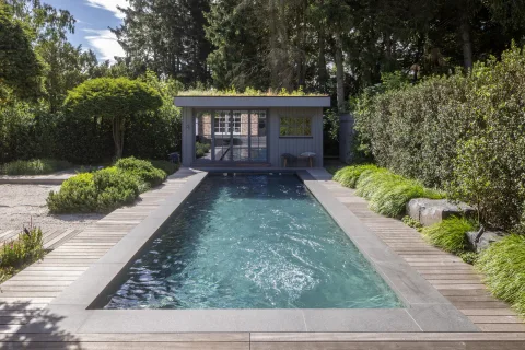 Becken mit Holzdeck, dahinter ein Poolhaus mit begrüntem Dach, Gräser und Kiesfläche