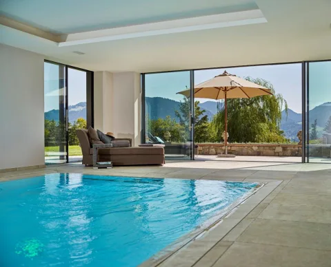 Innenbecken vor raumhoher Glasfront mit Blick auf Terrasse, Sonnenschirm und Bergkette