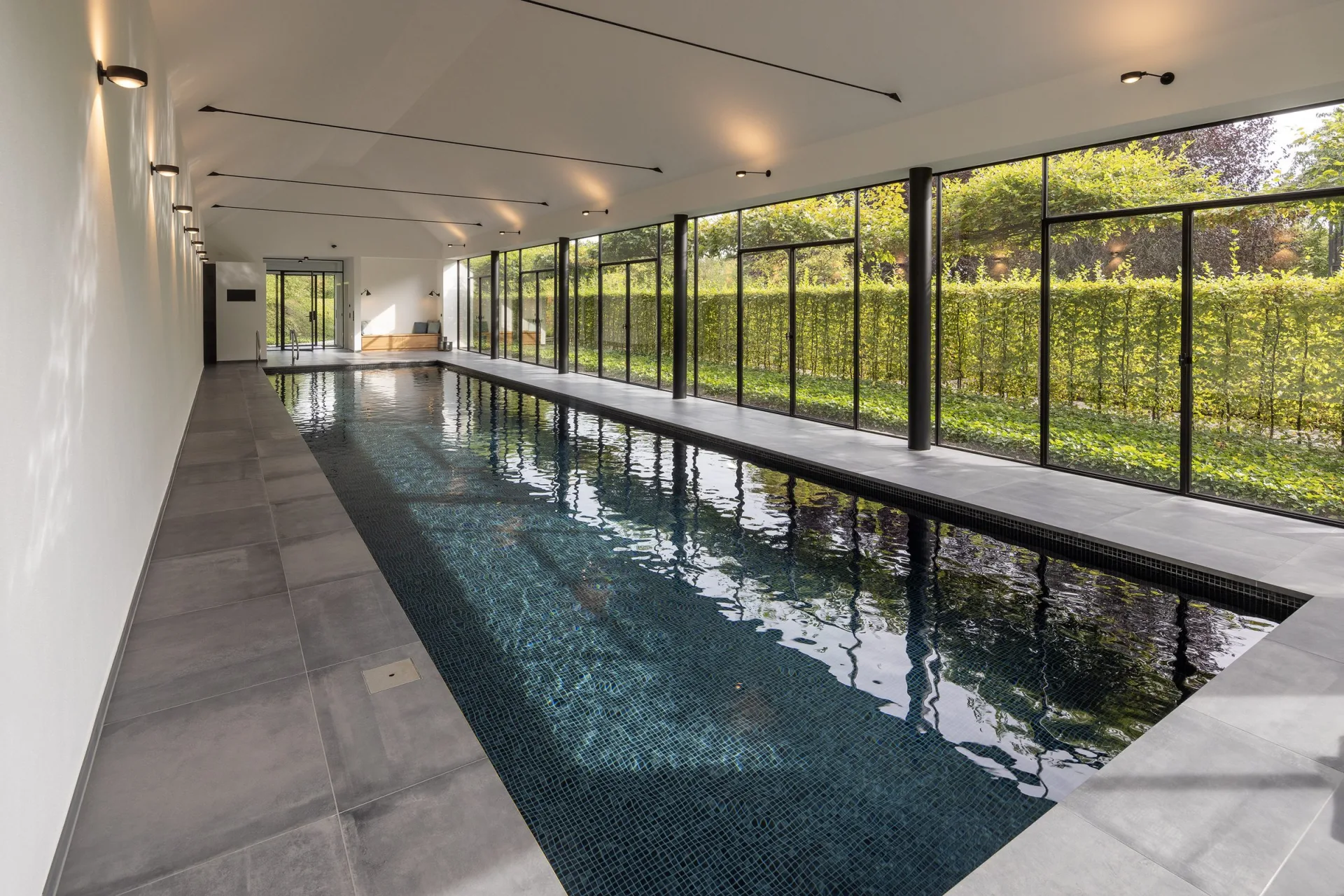 Innenpool als langes Schwimmbecken mit dunklem Mosaik, raumhohe Verglasung mit Blick in den Garten